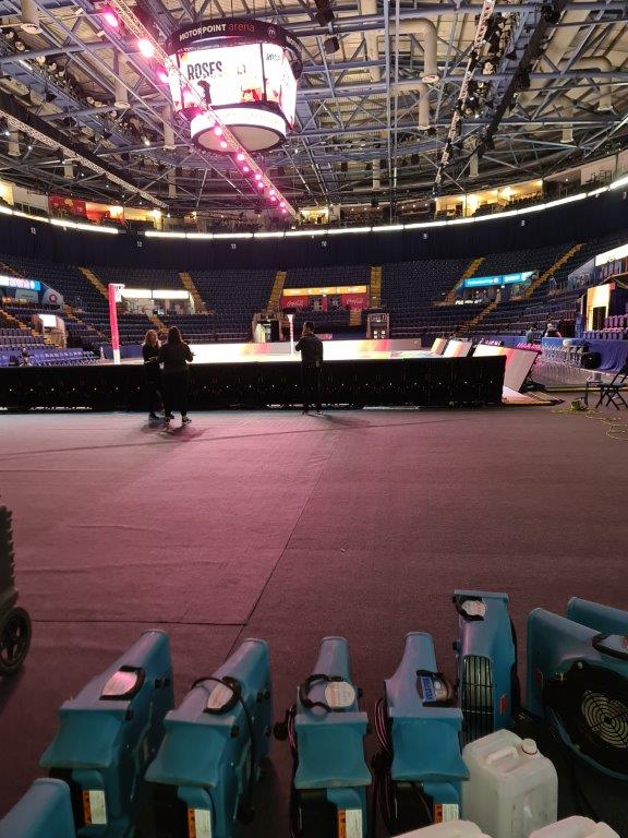 A view of the empty Motorpoint Arena, showing the performance stage with lighting, crew members preparing, and air movers positioned on the floor.